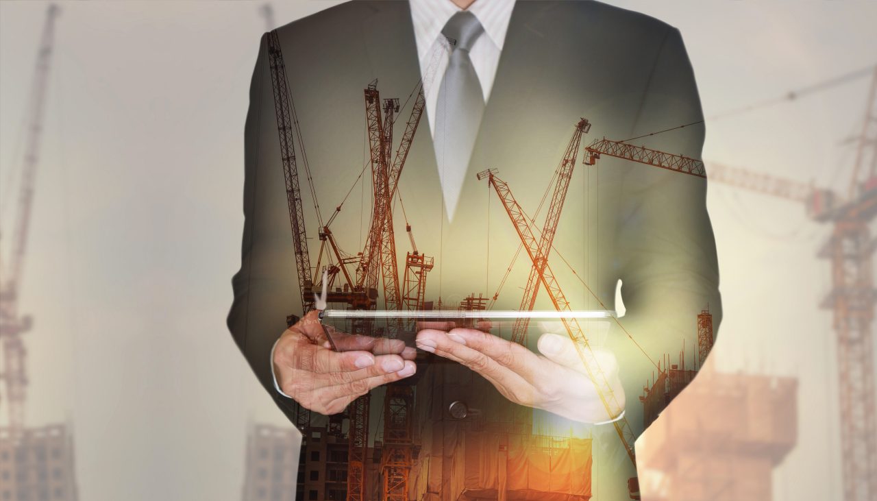 665721386 Double exposure of businessman hand hold tablet, construction crane, building and sunset in the evening as business, technology, communication and industry concept.